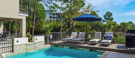 A swimming pool with lounge chairs and an umbrella, surrounded by trees and a building.
