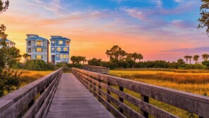 A wooden boardwalk leading to a building with multiple windows and balconies, surrounded by a natural landscape with trees and a sunset sky. - Boardwalk & Blue by Avantstay Sleeps 22 Infinity Pool + Elevator + Pool Table Steps to Sand (Hilton Head Island)