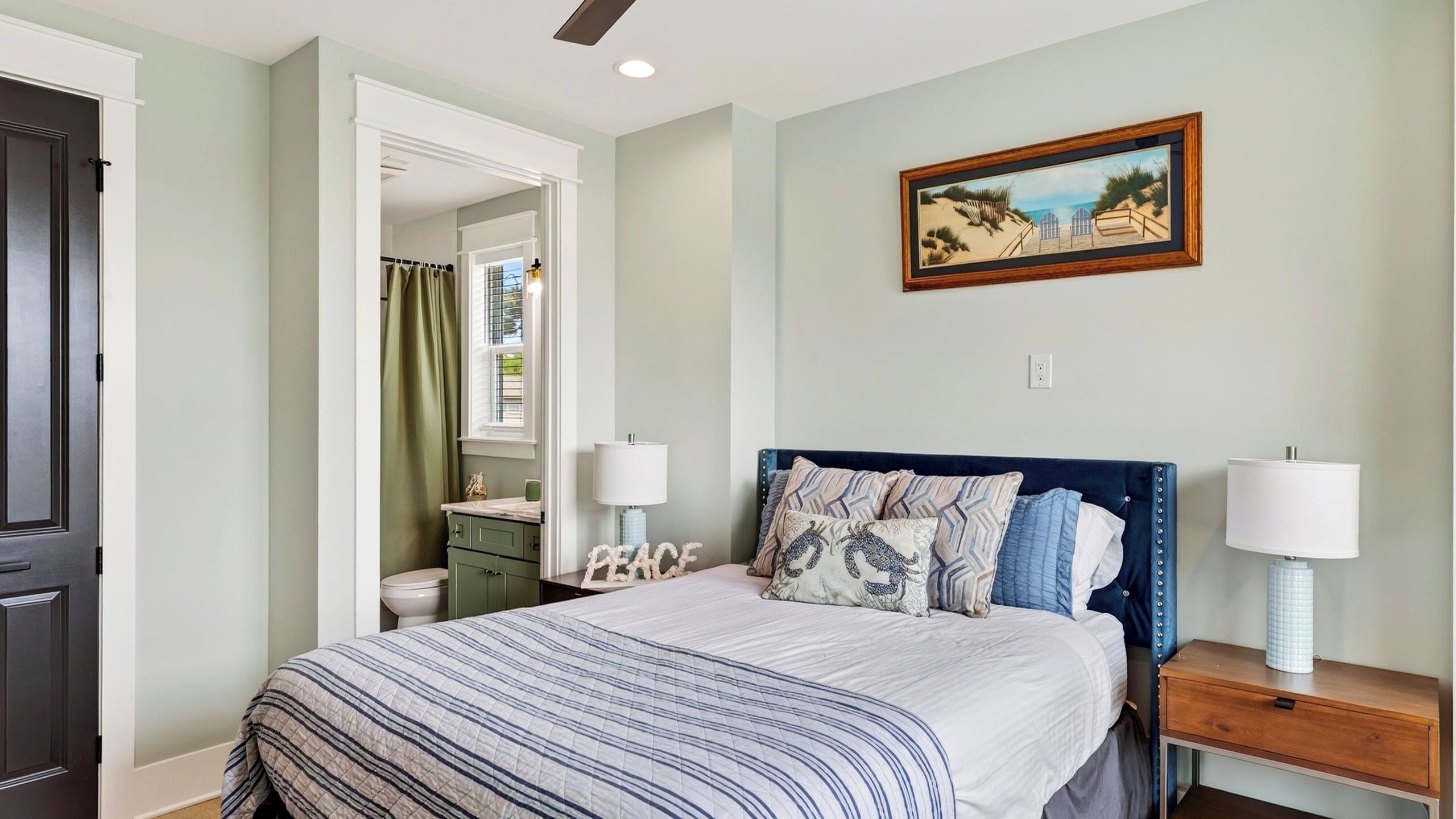 A bedroom with a bed, two bedside tables, a nightstand, a painting, and a bathroom visible through an open door.