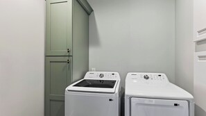A laundry room with two white front-loading washing machines and a light green cabinet. - Boardwalk & Blue by Avantstay Sleeps 22 Infinity Pool + Elevator + Pool Table Steps to Sand (Hilton Head Island)