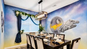 Un comedor con un mural de temática espacial, una araña y una mesa dispuesta para seis comensales.