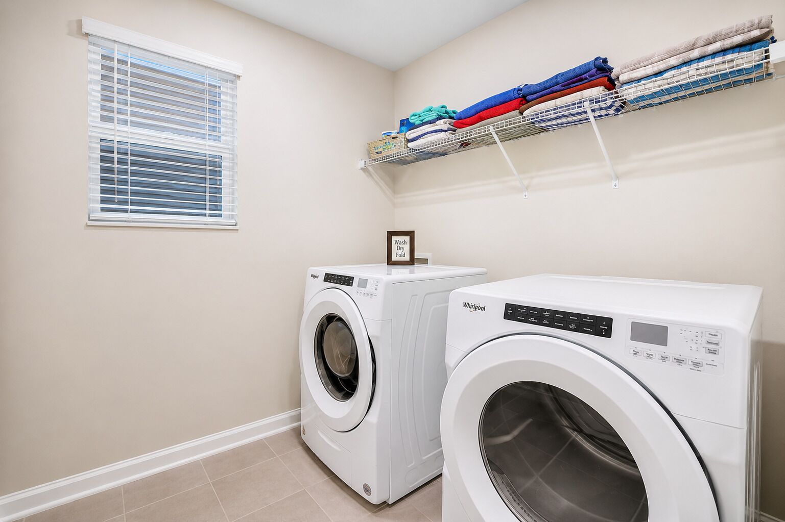 A laundry room with two front-loading washing machines, a window with blinds, and a shelf with folded towels.