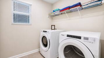 A laundry room with two front-loading washing machines, a window with blinds, and a shelf with folded towels.