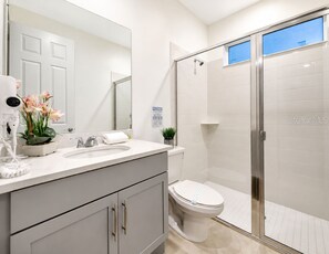 A bathroom with a white sink, a toilet, a shower with a glass door, and a mirror. - Suncrest Residence (Kissimmee)