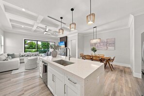 A modern kitchen with a central island, pendant lighting, and a dining area with a view of the outdoors. - New! Dream Chaser • New Luxury Build • Heated Saltwater Pool/Spa • 1 Mile to Vanderbilt Beach! (Naples)