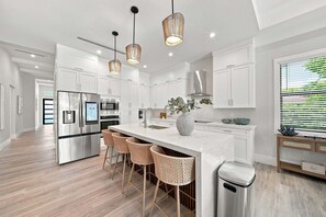 A modern kitchen with stainless steel appliances, a central island with bar stools, pendant lighting, and a view of greenery outside. - New! Dream Chaser • New Luxury Build • Heated Saltwater Pool/Spa • 1 Mile to Vanderbilt Beach! (Naples)