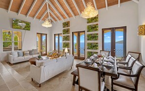 A spacious living area with a dining table set for four, a sofa, and a view of the sea through large windows. - 3 King Suites | Pool + Mahogany Run | Magens Bay (St. Thomas)