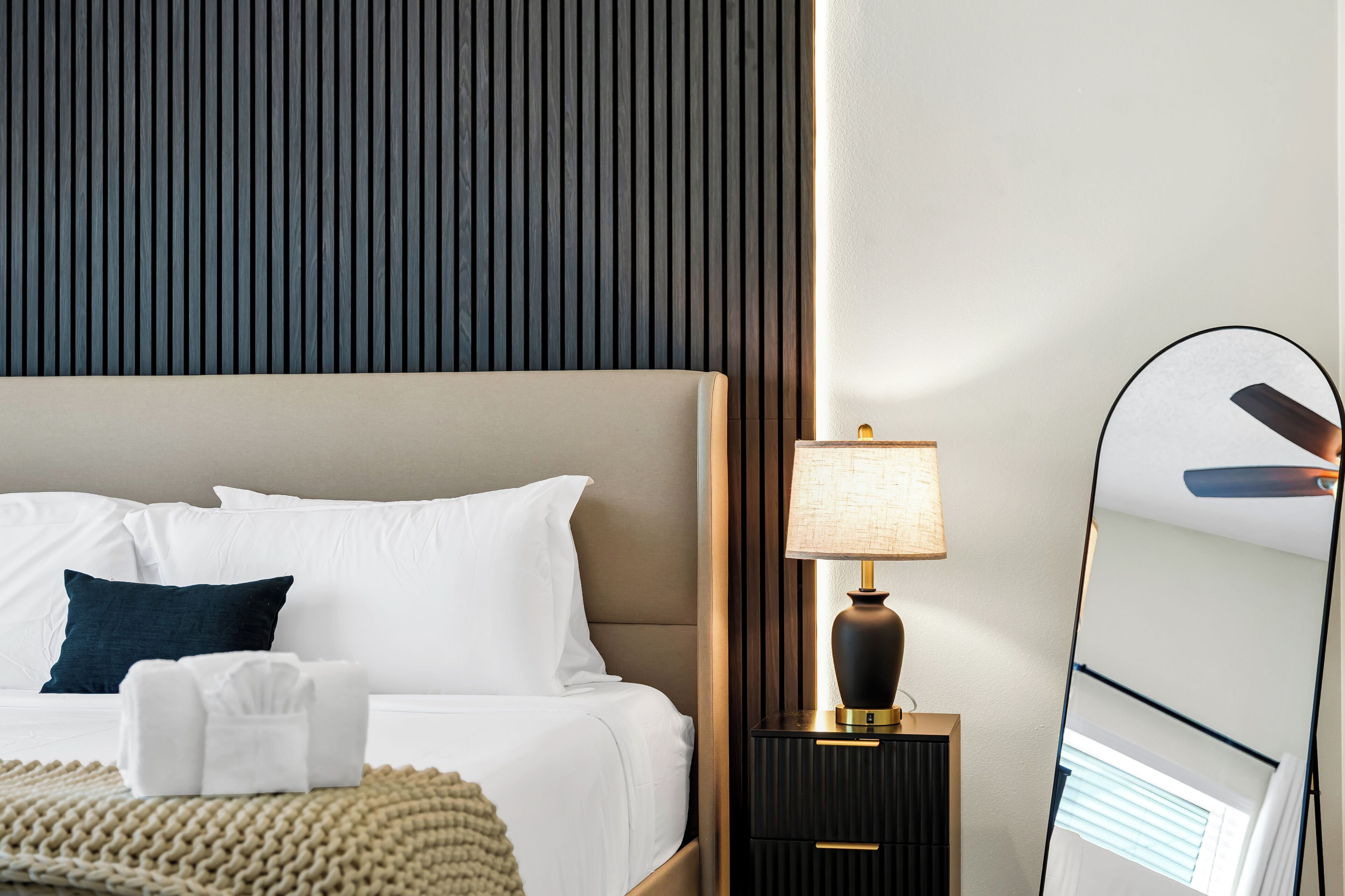 A neatly made bed with white and dark pillows, a bedside lamp, a small wooden nightstand, and a mirror reflecting part of the room.
