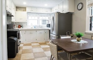Een kleine keuken met witte kastjes, een roestvrijstalen koelkast en een eettafel met stoelen.