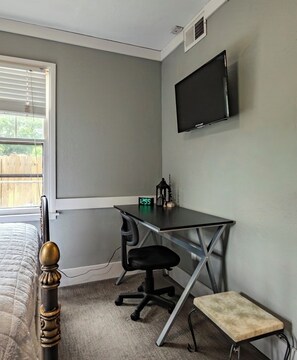 Un dormitorio con cama, un escritorio y una silla, un taburete pequeño, un televisor montado en la pared y una ventana con persianas.