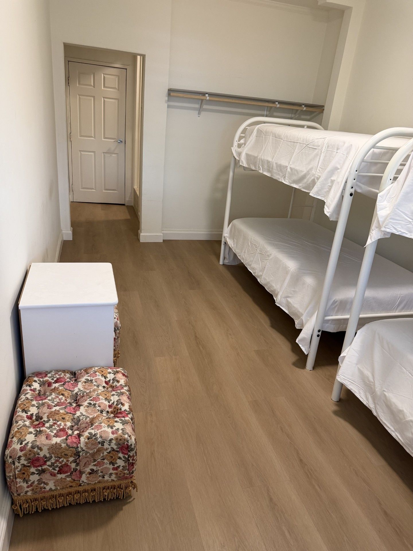A room with a bunk bed, a small white chest of drawers, and a floral-patterned ottoman.