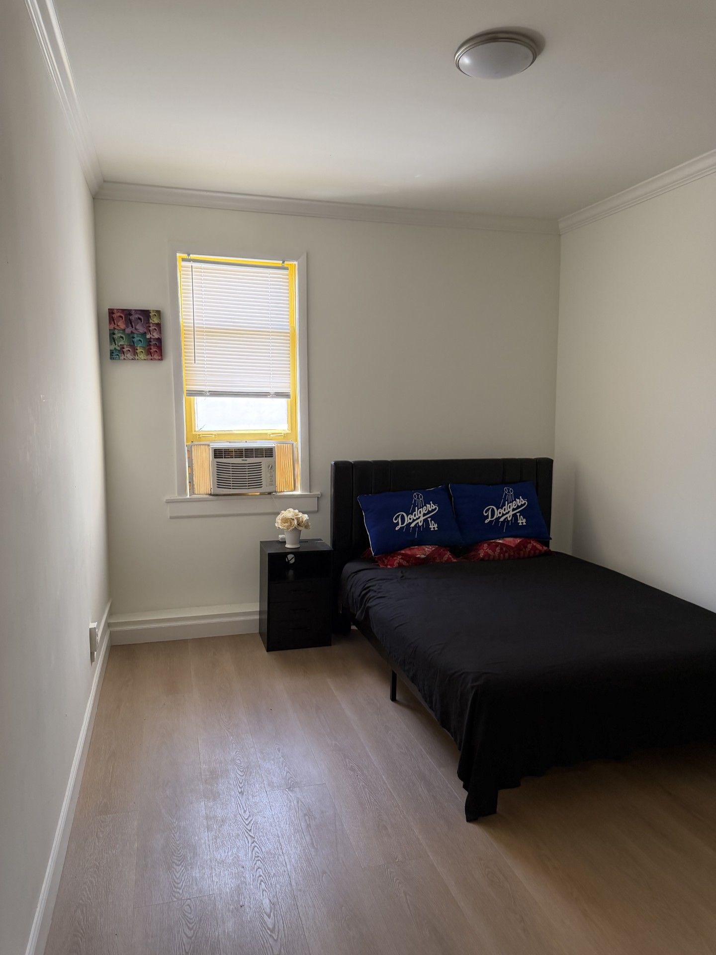 Une chambre à coucher avec un lit, une table de chevet, une fenêtre avec des stores et un petit objet décoratif sur la table de chevet.