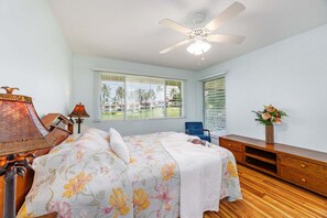 Ein Schlafzimmer mit einem Bett, einem Deckenventilator, einem Nachttisch, einer Lampe, einem Sessel, einer Kommode und einem Fenster mit Jalousien.