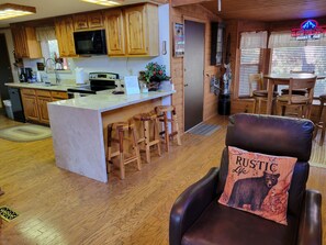 A cozy interior with a kitchen featuring wooden cabinets, a central island with stools, and a leather chair with a bear pillow. - Cozy Cabin between the Pines (Payson)