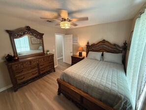 Un dormitorio con cama de madera, cómoda, espejo, ventilador de techo y puerta que da a otra habitación.