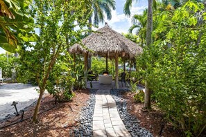 A thatched-roof gazebo surrounded by lush greenery and a stone pathway. - Coastal Beauty - Canal Front, Dock, Pool, Spa, Game Room by Anna Maria Life Vacation Rentals (Anna Maria)