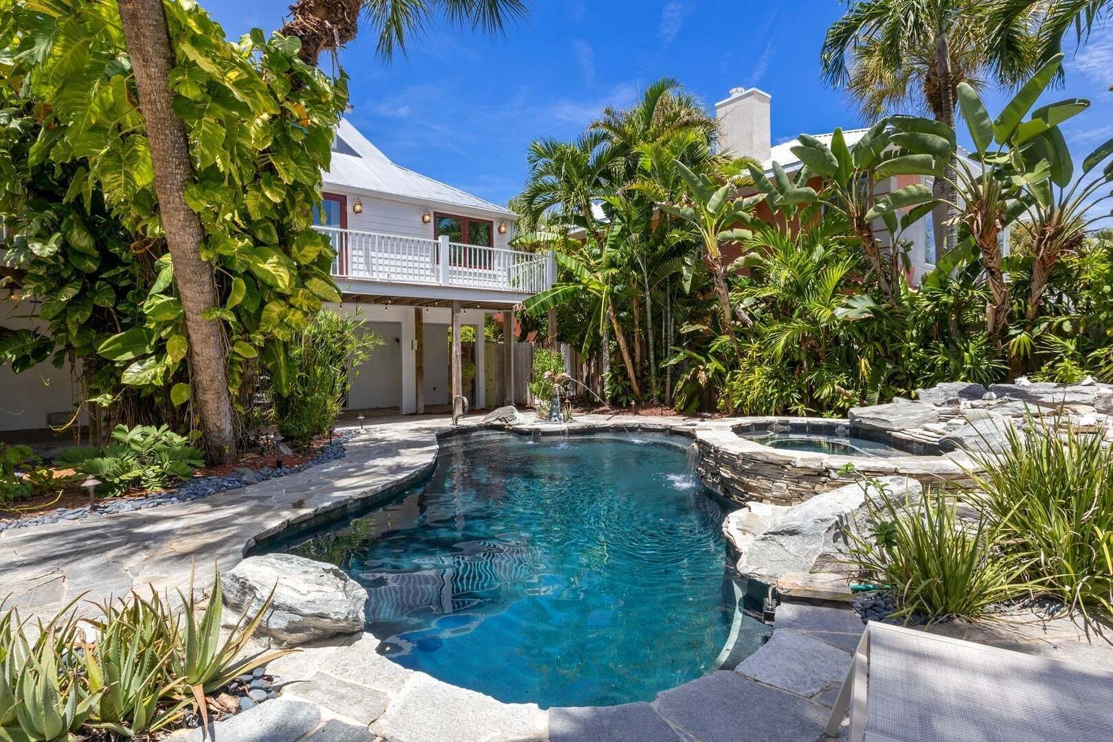 A pool with a stone edge and a small waterfall feature, surrounded by tropical vegetation and a building with a balcony.