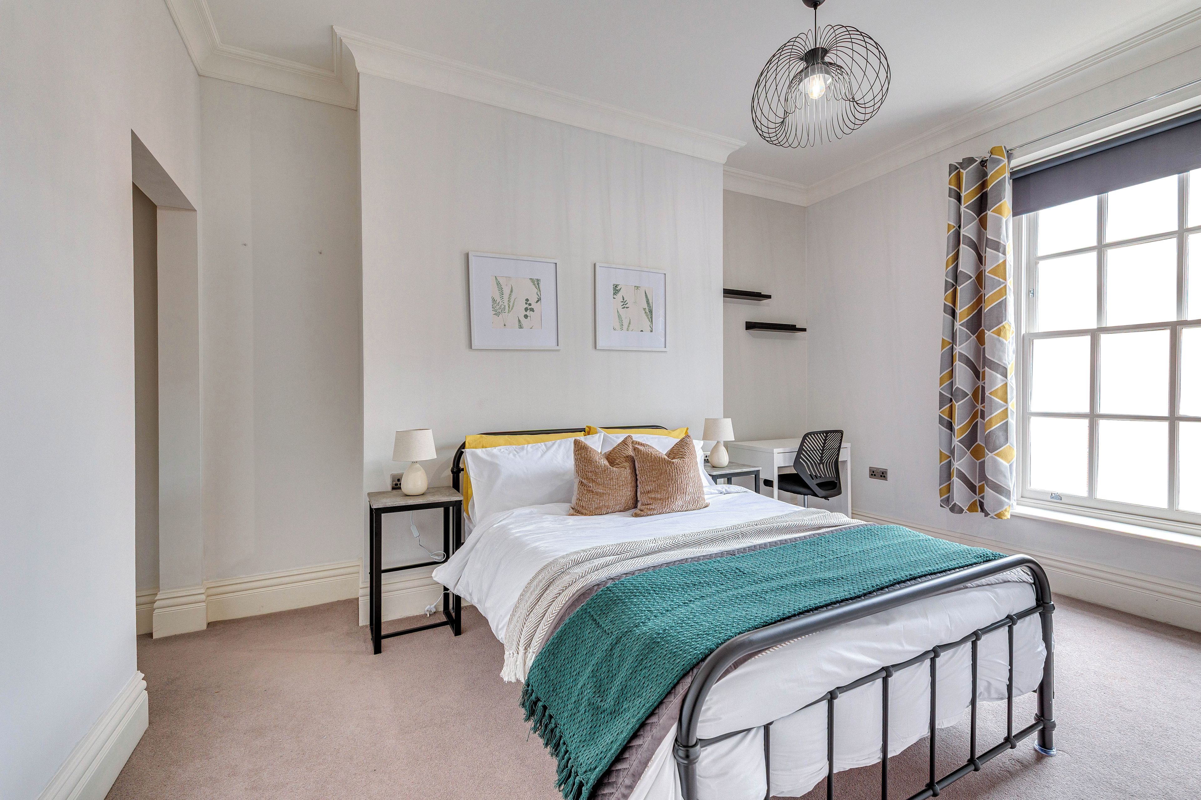 A bedroom with a bed, bedside tables, a desk, a chair, a window with curtains, and two wall-mounted pictures.