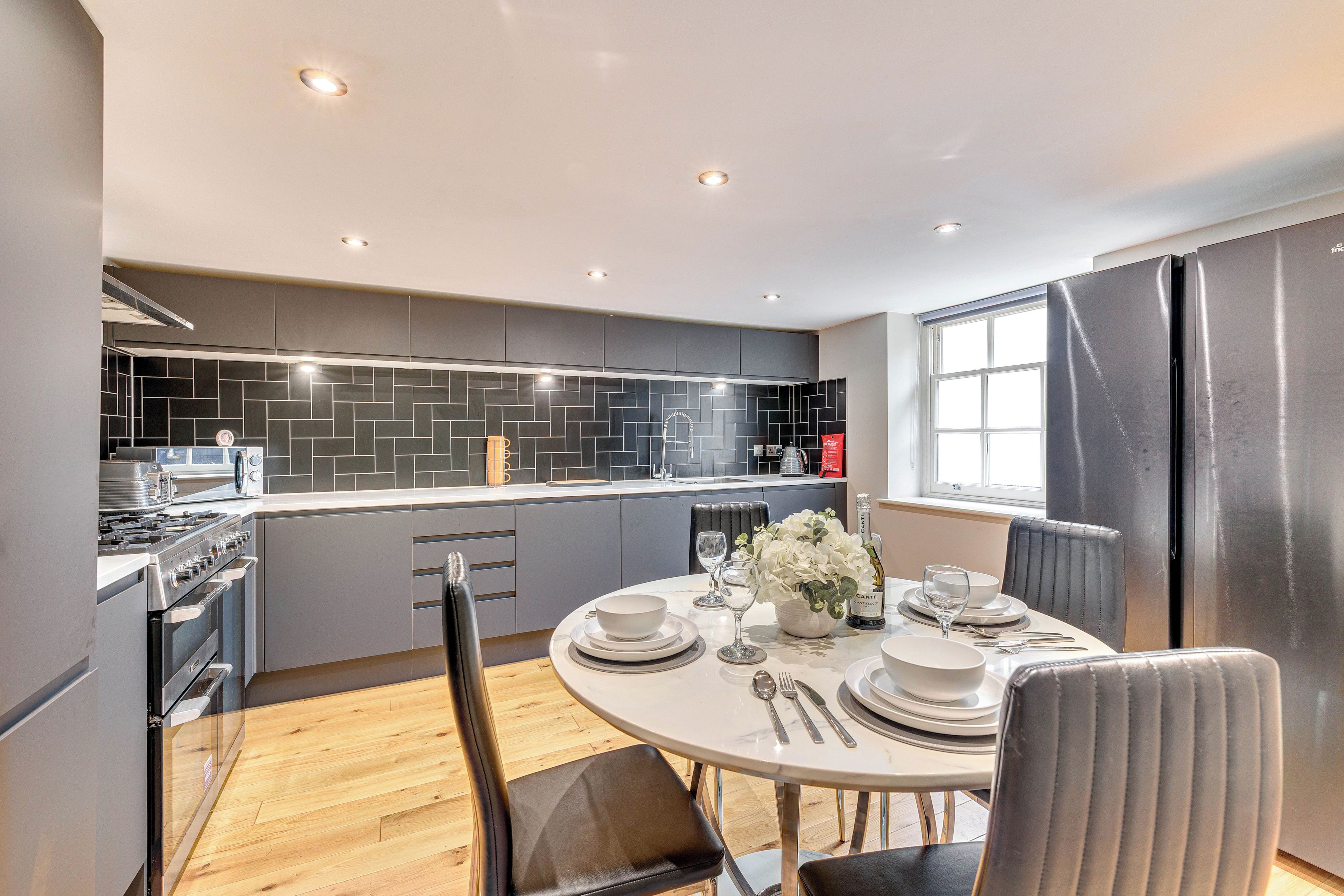 A modern kitchen with a dining area, featuring a large refrigerator, stove, and black tiled backsplash.