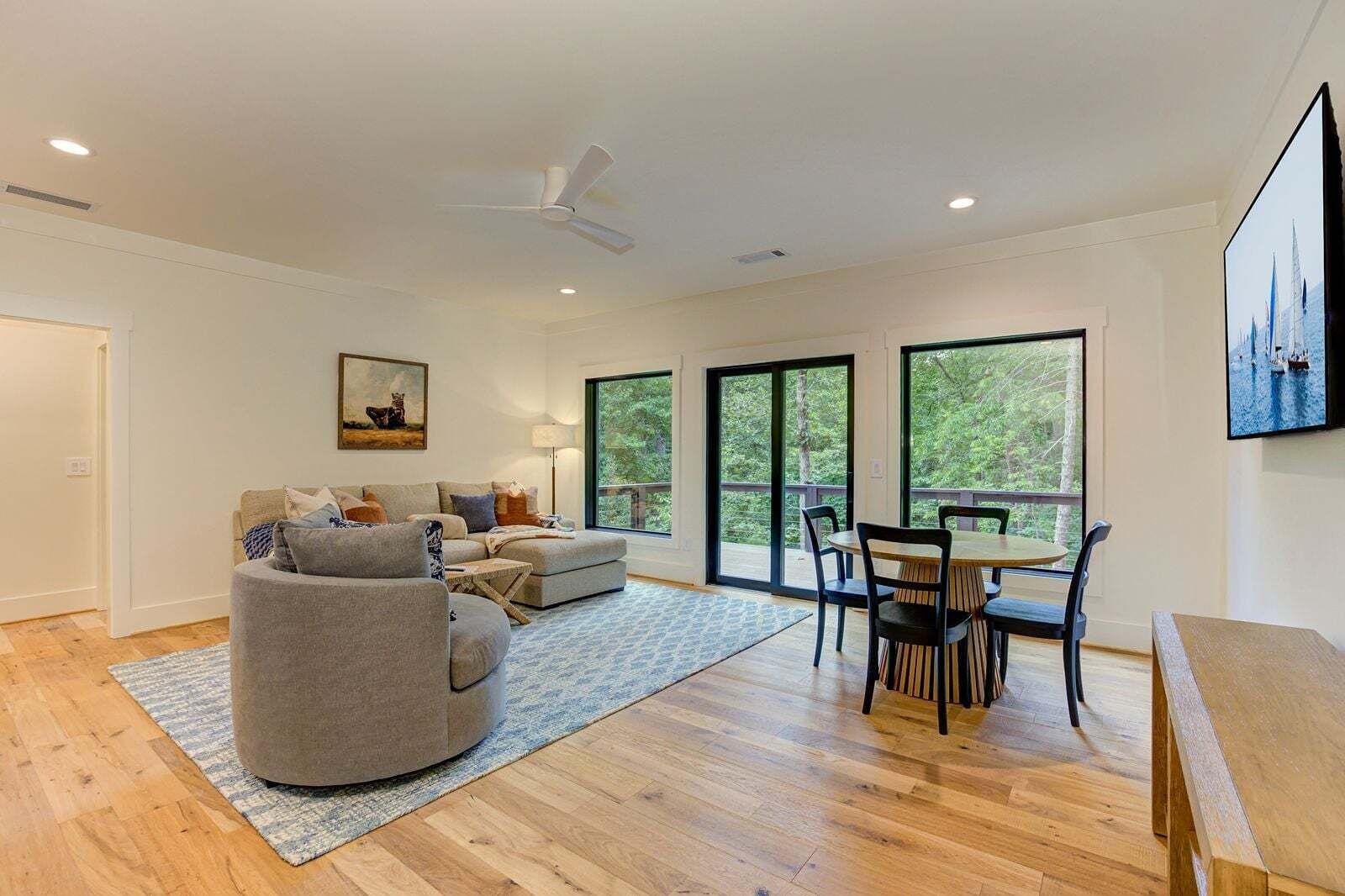 Una sala de estar moderna con un sofá, una mesa de comedor con sillas y ventanas amplias que ofrecen vista a árboles.