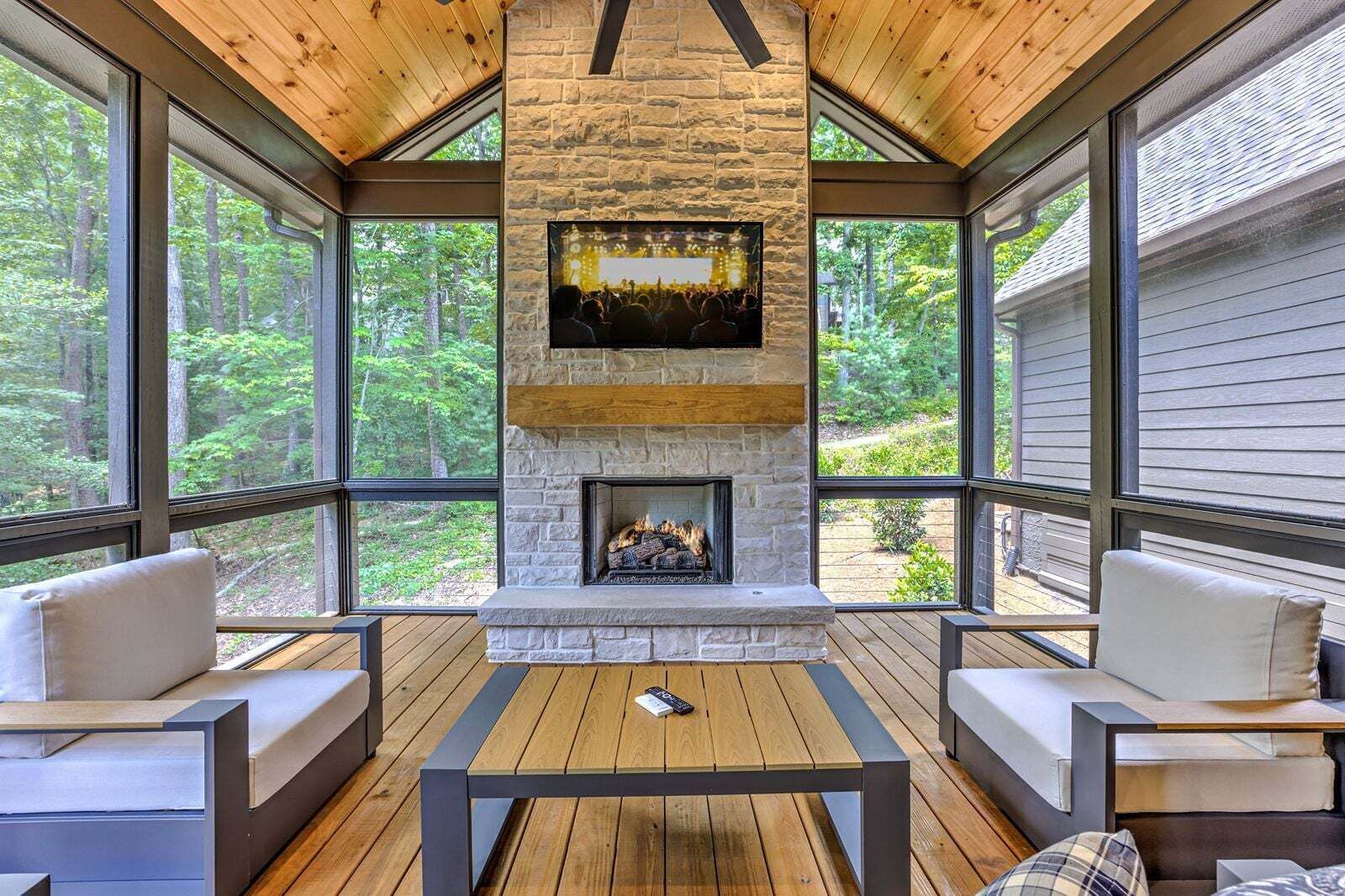 A cozy room with a stone fireplace, wooden ceiling, and large windows overlooking a forest.