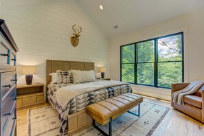 Ein Schlafzimmer mit einem großen Bett, einem Holzkopfteil, einer Hirschdekoration, einer Bank, einem Ledersofa und einem großen Fenster mit Blick auf Bäume.