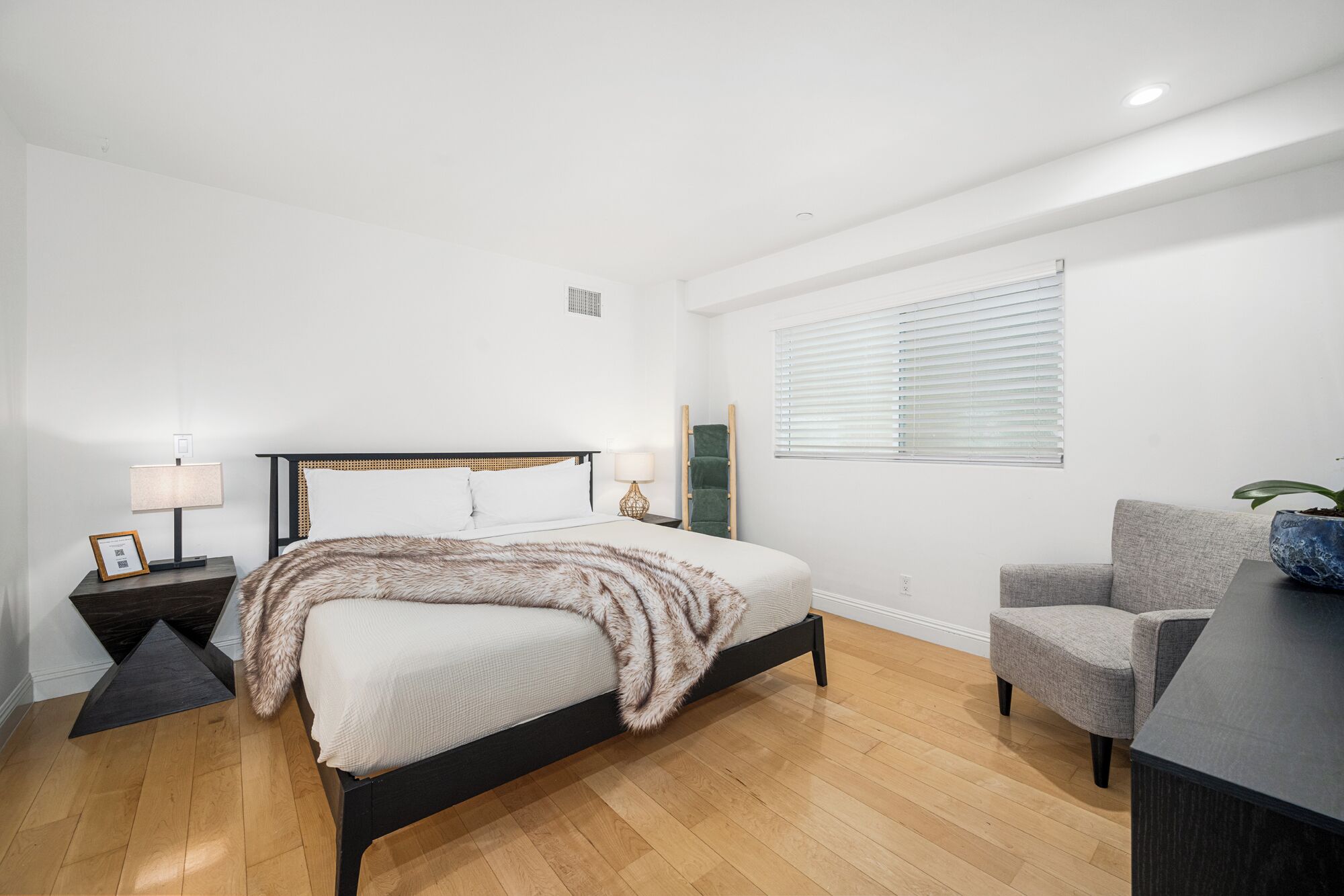 A bedroom with a bed, bedside tables, a chair, a window with blinds, and a plant.