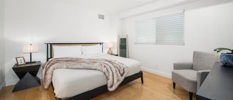 A bedroom with a bed, bedside tables, a chair, a window with blinds, and a plant.