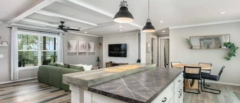 A modern kitchen with a marble countertop, a dining area with a round table and chairs, and a living room with a green sofa.
