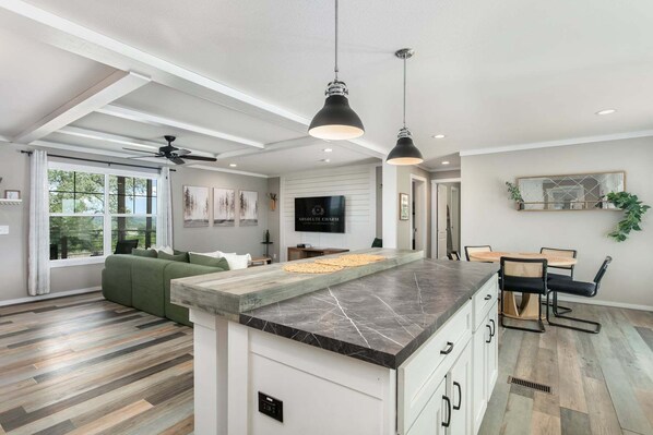 A modern kitchen with a marble countertop, a dining area with a round table and chairs, and a living room with a green sofa.