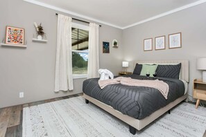 Ein Schlafzimmer mit Bett, einem Fenster mit Vorhängen, einem Nachttisch mit Lampe und Wandbildern.