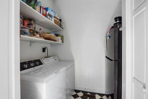 A compact laundry room with a washing machine, a refrigerator, and shelves stocked with various food items. - The Ark - Add to the History and Memories of Sullivan (Sullivans Island)