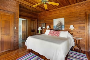 A wooden-paneled room with a bed, a ceiling fan, and a painting on the wall. - The Ark - Add to the History and Memories of Sullivan (Sullivans Island)
