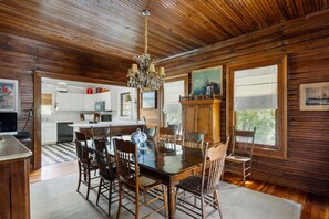 A wooden-paneled dining room with a large table and chairs, a chandelier, and a view of the kitchen. - The Ark - Add to the History and Memories of Sullivan (Sullivans Island)