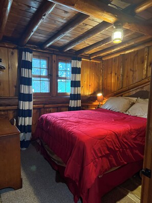 A wooden cabin bedroom with a bed, a nightstand, a lamp, and a window with curtains. - GORGEOUS Lakefront Log Home 3-bedrooms tucked into serene woods by Gaylord (Gaylord)