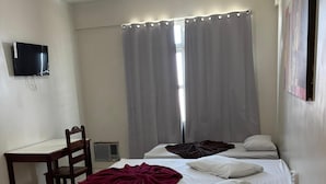 Ein Hotelzimmer mit Bett, Schreibtisch, Stuhl, einem an der Wand befestigten Fernseher und einem Fenster mit Vorhängen.