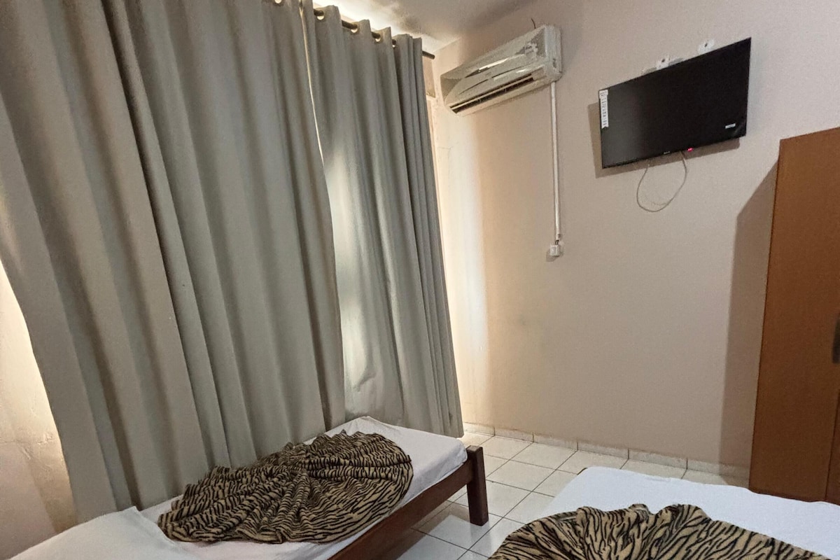 A room with two beds, one with a zebra-patterned blanket and another with a plain white sheet. There is a television mounted on the wall and an air conditioning unit above it.