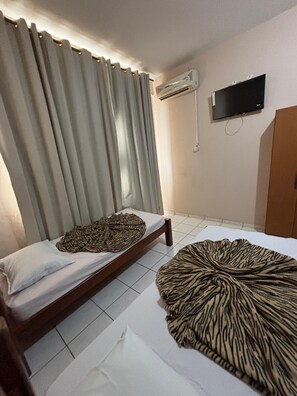 A room with two beds, one with a zebra-patterned blanket and another with a plain white sheet. There is a television mounted on the wall and an air conditioning unit above it. - Hotel Via Montes (Montes Claros)