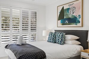 Une chambre à coucher avec un lit, des rideaux blancs, une lampe fixée au mur et un tableau abstrait encadré.