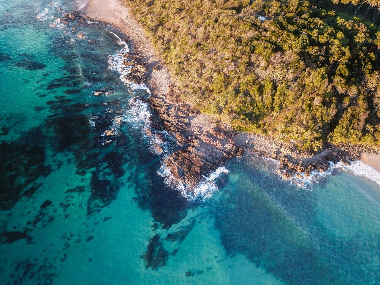 An aerial view of a coastal area with clear turquoise waters, rocky shoreline, and dense green vegetation.