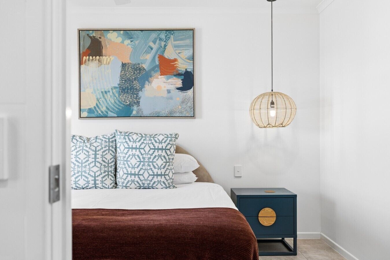Een slaapkamer met een bed, kussens, een nachtkastje, een aan de muur hangend kunstwerk en een hanglamp.