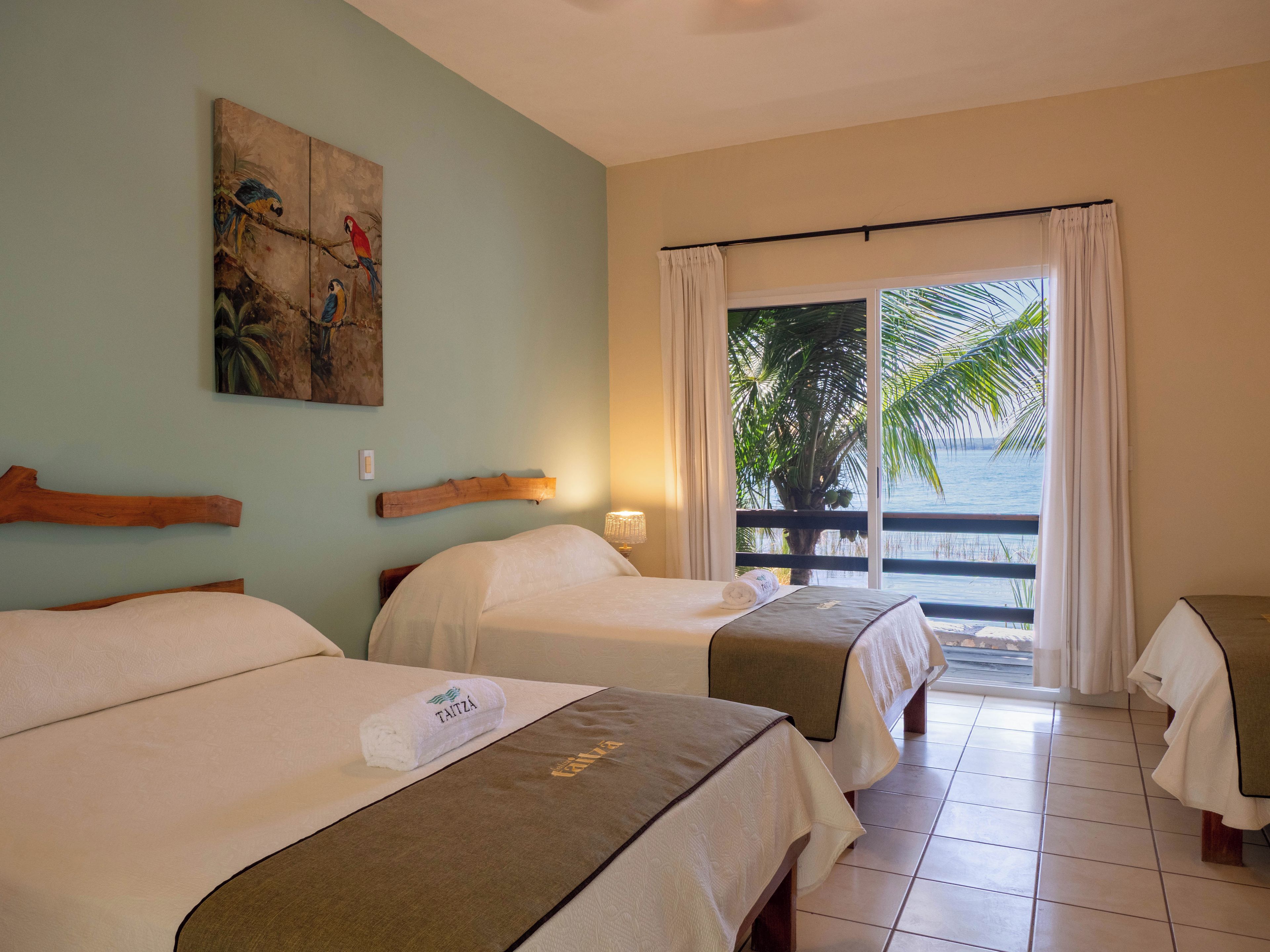 A hotel room with two beds, a painting on the wall, a view of the sea through a window, and a palm tree outside.
