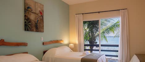 A hotel room with two beds, a painting on the wall, a view of the sea through a window, and a palm tree outside.