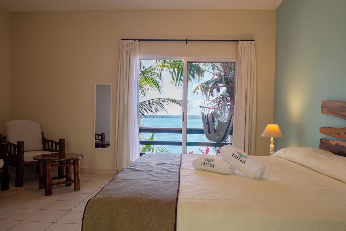 Habitación de hotel con una cama, una silla, una mesita pequeña, un sillón colgante y vista al océano a través de un ventanal grande.