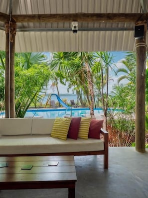 A covered seating area with a view of a pool and palm trees.