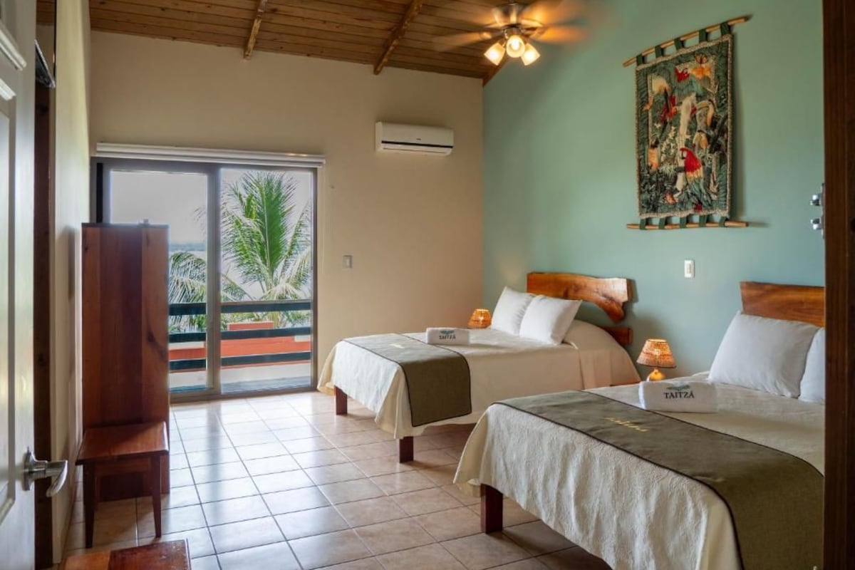 Habitación de hotel con dos camas, ventilador de techo, ventana grande con vistas y un banco de madera.