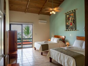 Habitación de hotel con dos camas, ventilador de techo, ventana grande con vistas y un banco de madera.