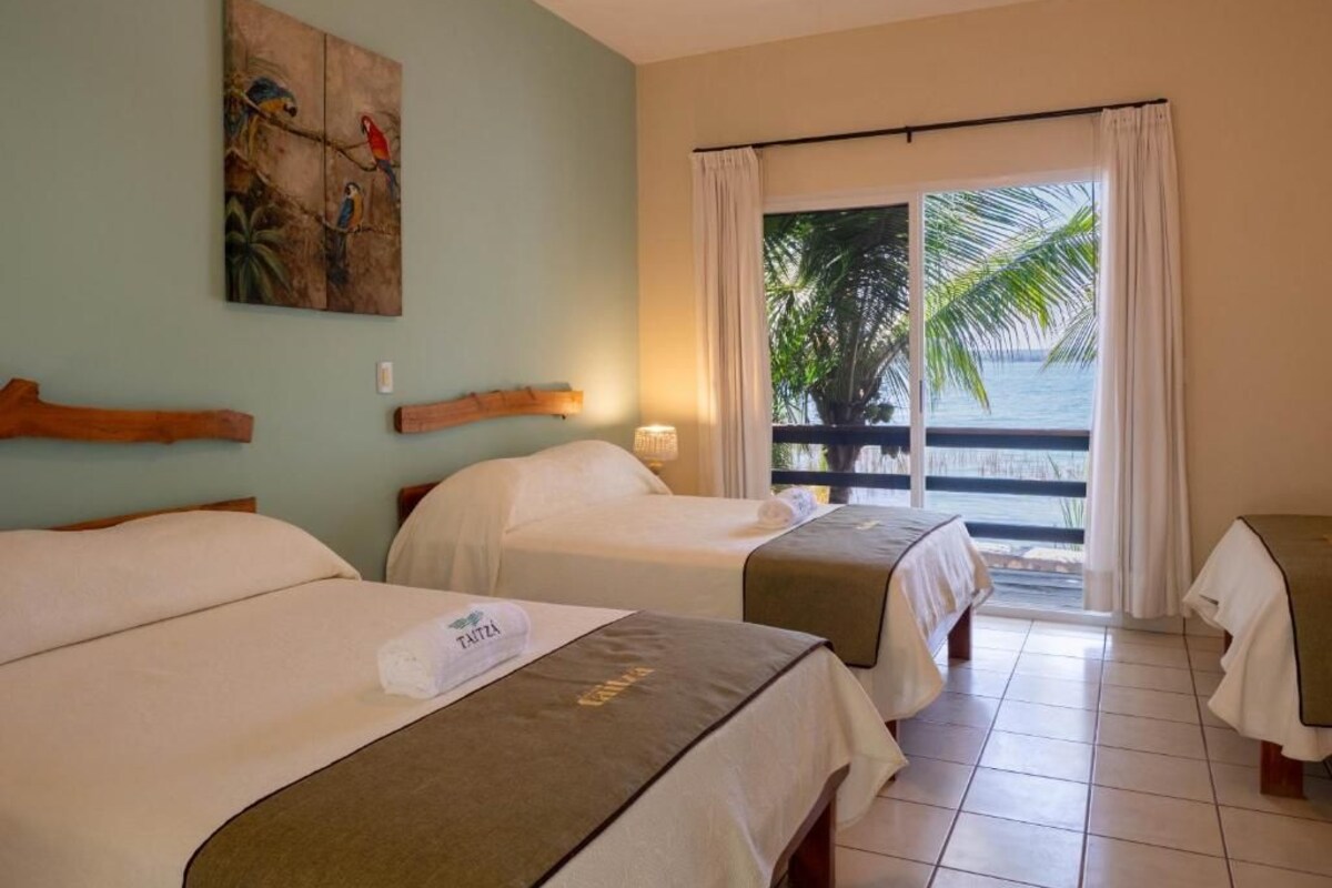 Habitación de hotel con dos camas, un cuadro en la pared, vista al océano a través de una puerta corredera y un balcón con una palmera.