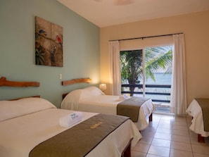 Habitación de hotel con dos camas, un cuadro en la pared, vista al océano a través de una puerta corredera y un balcón con una palmera.