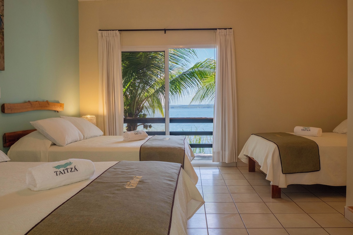 Habitación de hotel con dos camas, un balcón con vista a palmeras y al mar, y un cabecero de madera.